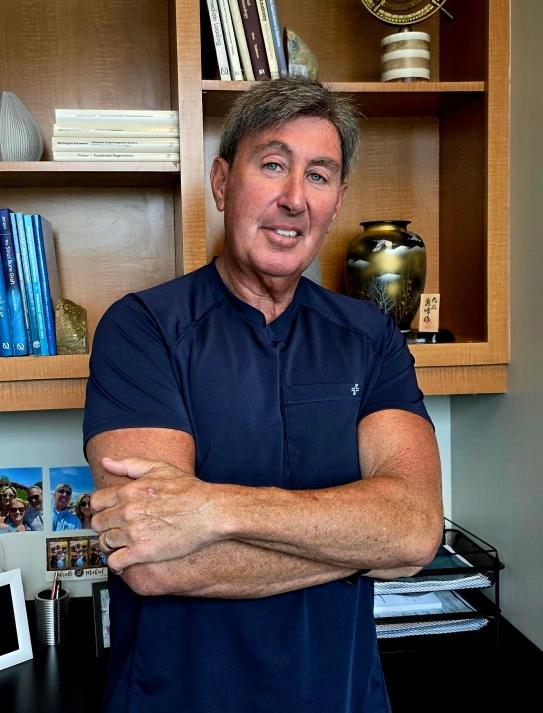 A medium portrait of a middle-aged male professional with short, textured greyish-brown hair and blue eyes, smiling with his arms crossed. He is wearing a short-sleeved dark navy blue scrub top. He stands in an office setting in front of a wooden bookshelf lined with books, a decorative vase, and small personal photos pinned to the side wall.