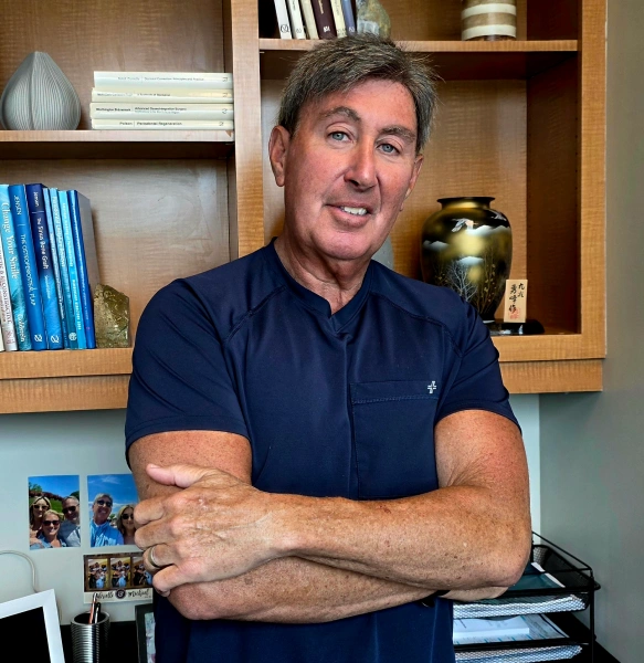 A medium portrait of a middle-aged male professional with short greyish-brown hair, smiling with his arms crossed in a dark navy blue medical scrub top. He is standing in front of a light wood bookshelf containing medical books and decorative items, with family photographs visible on the left wall background.